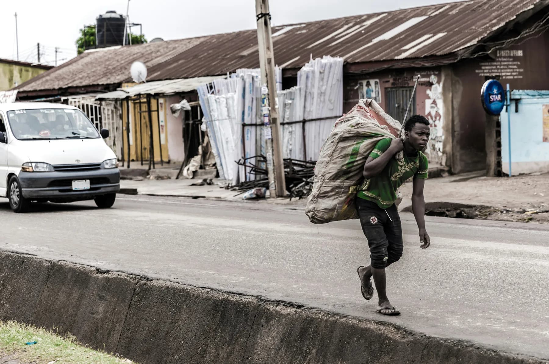 A young man carrying a full sack along a roadside past a row of shops
