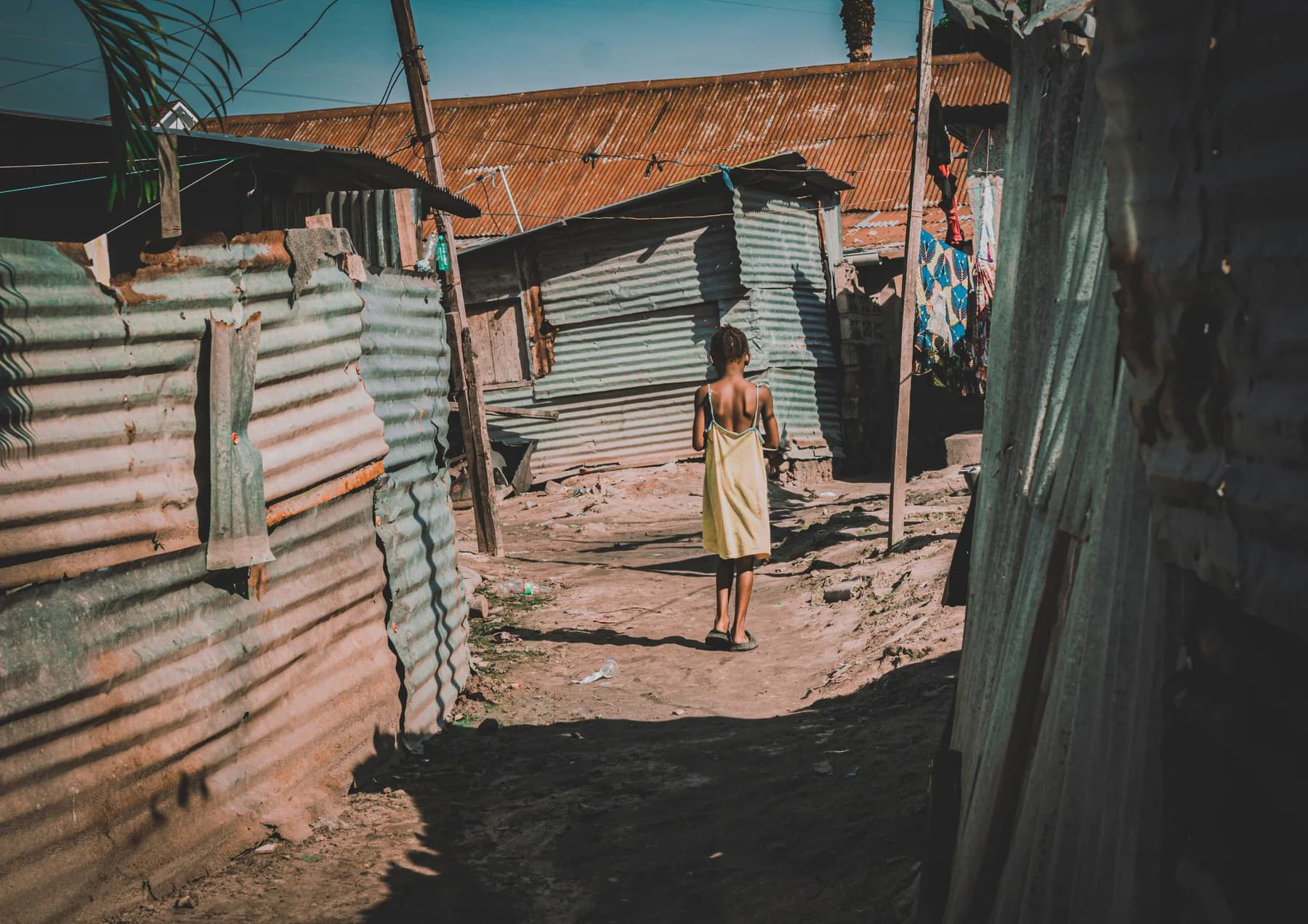 A girl walking alone down a narrow lane between corrugated-iron homes