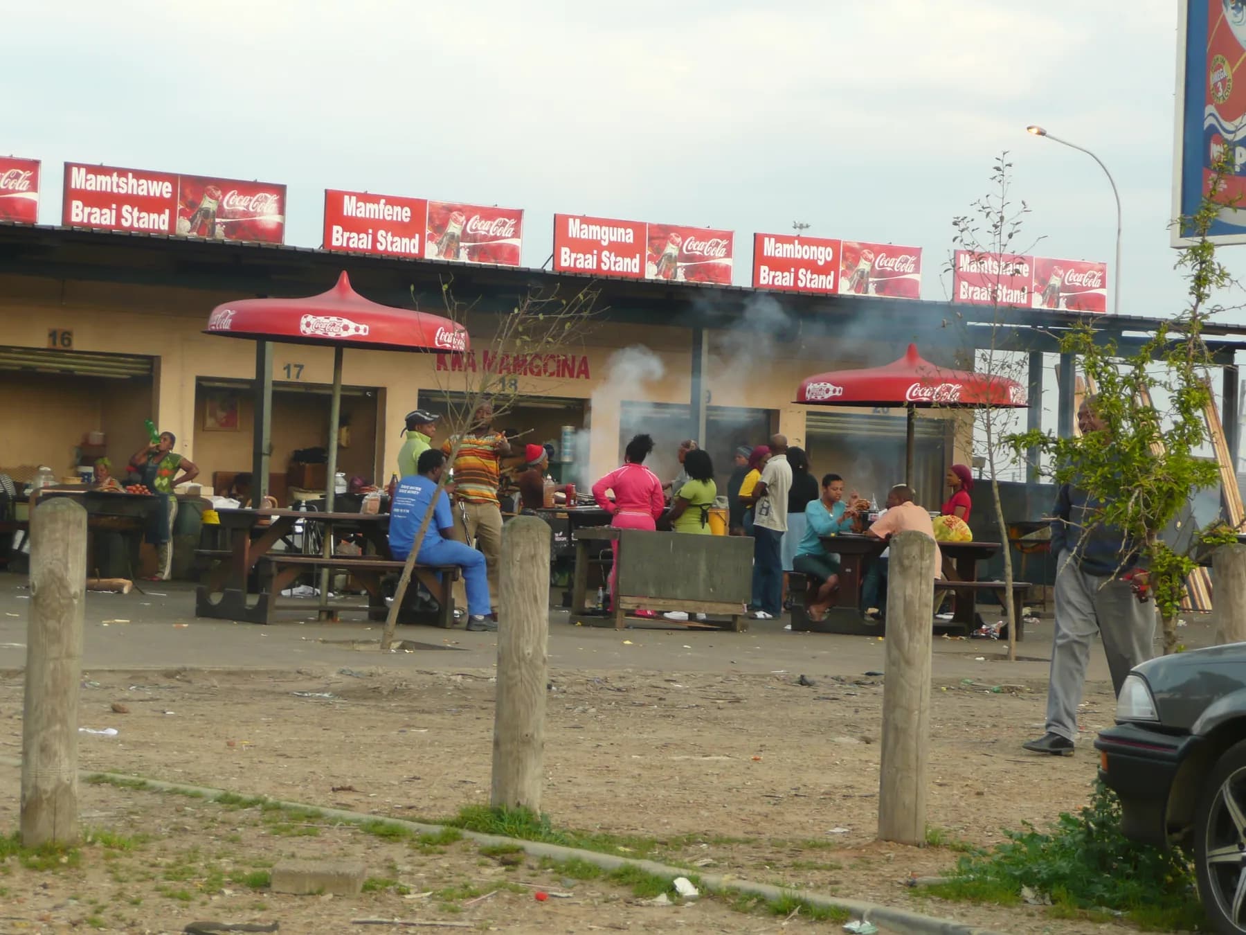 People eating together at a row of township braai stands, smoke drifting over them