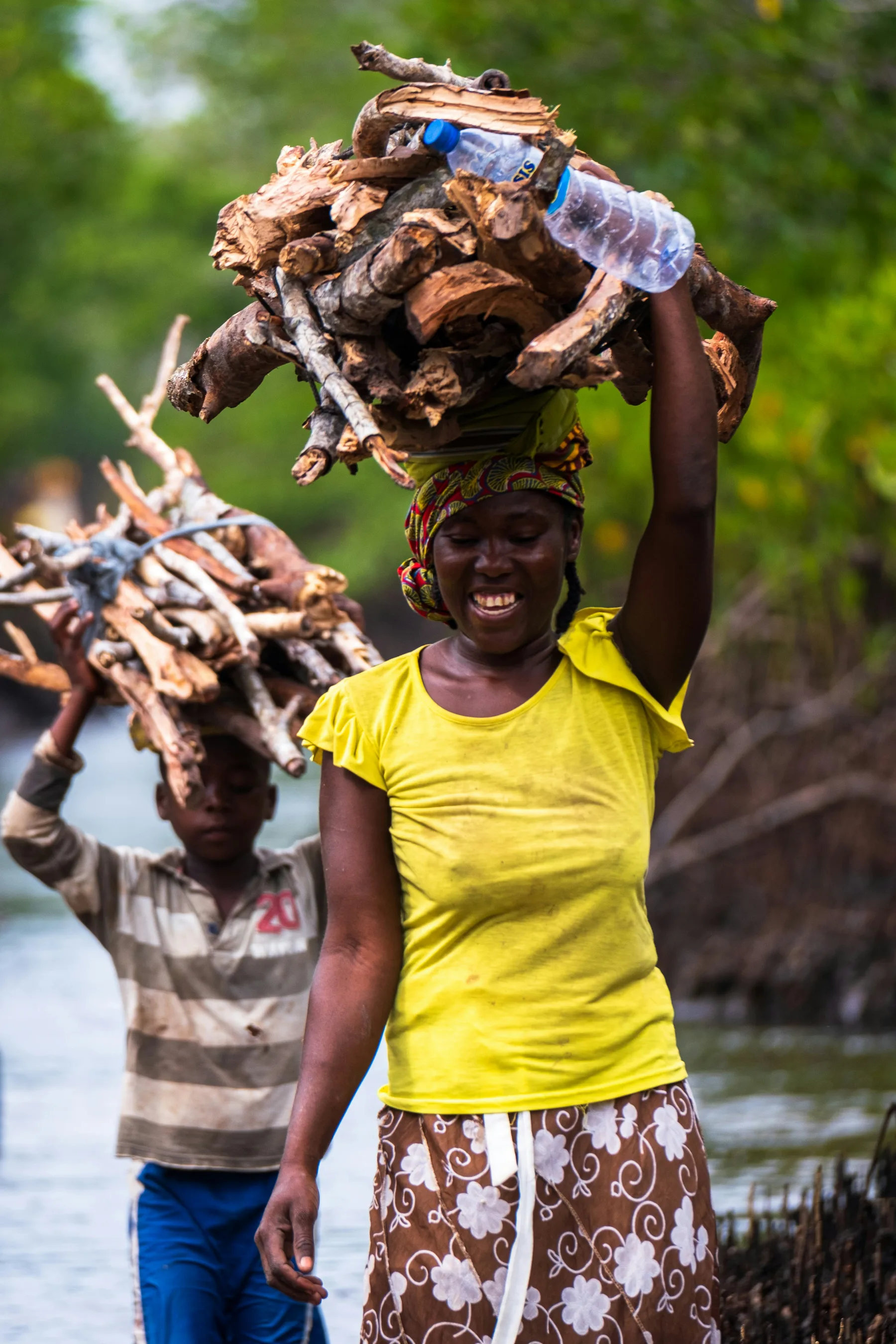 A woman and a boy carrying firewood home along a waterside path