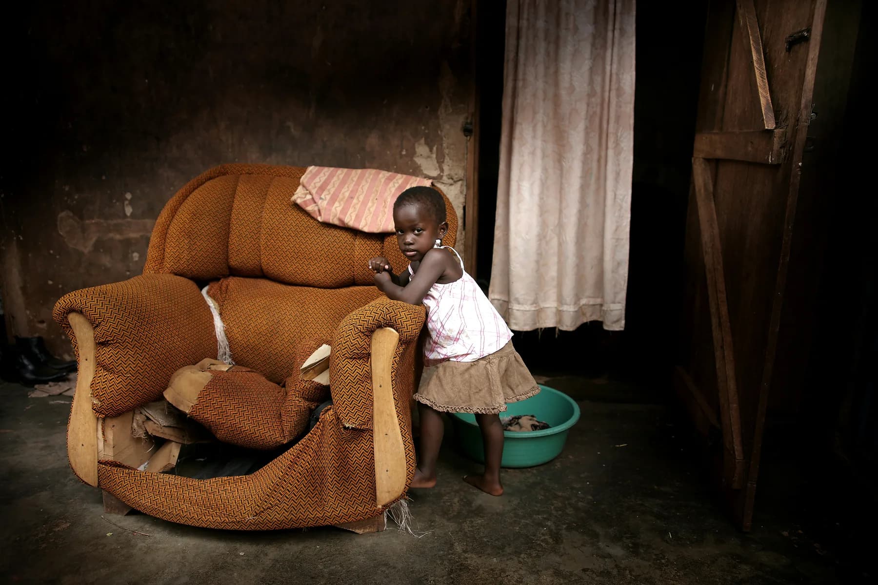 A small girl standing beside a worn armchair in a dim room